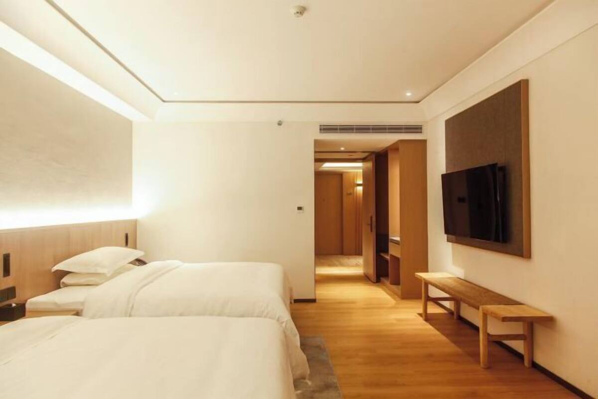 A hotel room with two beds, a bench, a TV, and a hallway leading to another room.