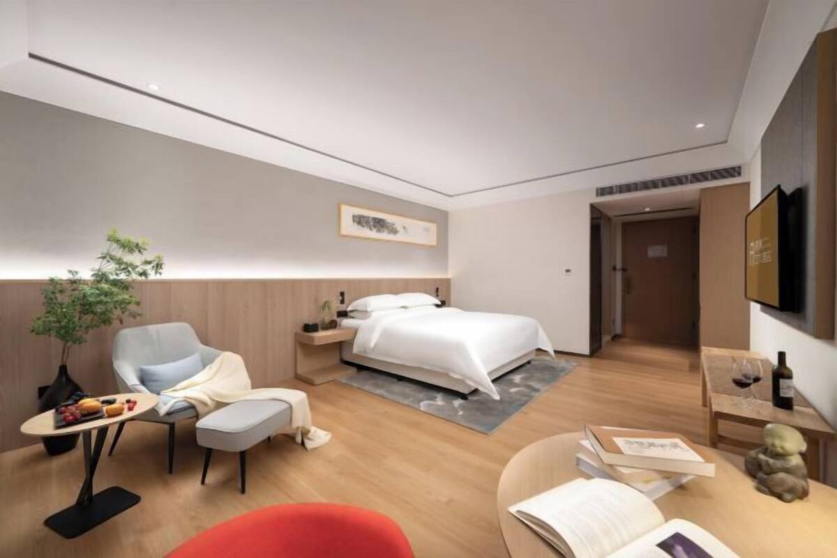 A modern hotel room with a bed, a desk, a chair, a small table with a plant, and a bookshelf.