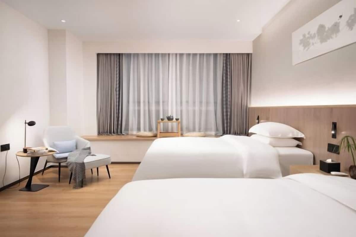 A hotel room with a large bed, a smaller bed, a desk with a chair, a window with curtains, and a wall-mounted artwork.
