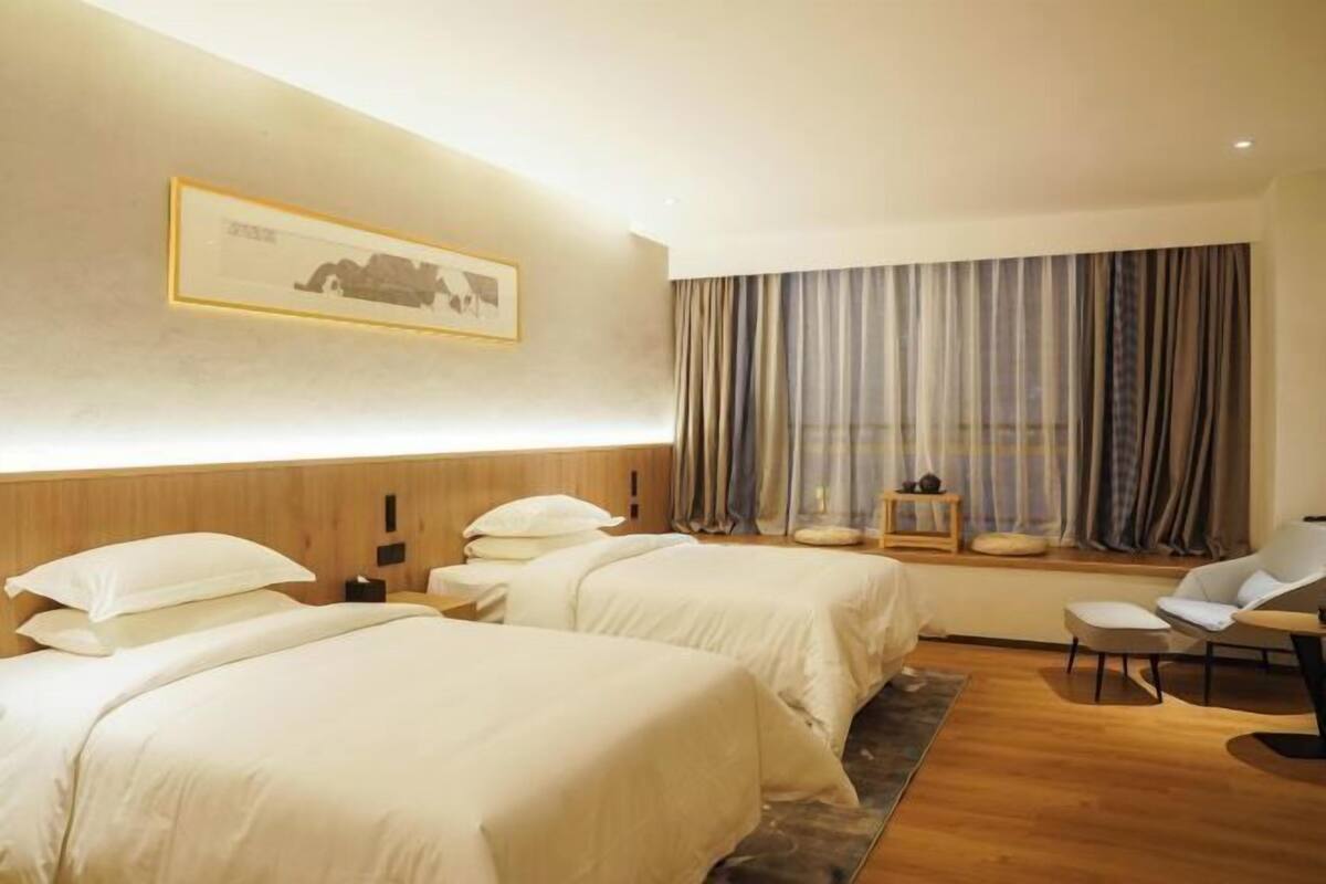 A hotel room with two beds, a desk, a chair, a window with curtains, and a framed picture on the wall.