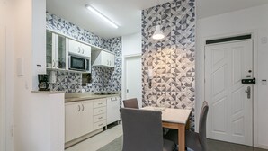 Kompakti keittiö, jossa on valkoiset kaapit, mikroaaltouuni ja kahvinkeitin. Keittiössä on ruokapöytä, jossa on kaksi tuolia, ja pöydän takana on kuvioitu seinä.