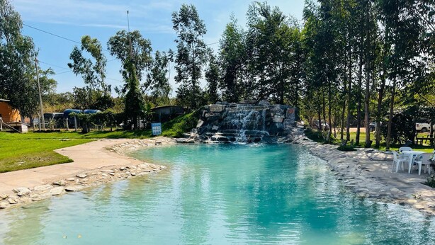 Piscina cristalina con una cascada, rodeada de árboles y un área de picnic con sillas blancas.