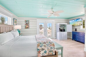 A bedroom with a bed, a ceiling fan, a TV, a kitchen area with cabinets, and a view through the door. - Two Houses from Beach! Rooftop, Pool & Game Room (Holmes Beach)
