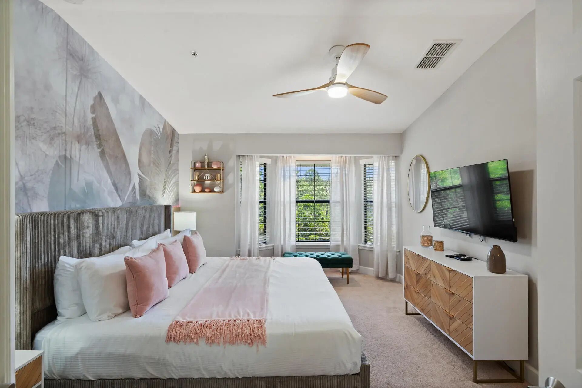 Un dormitorio con una cama grande, ventilador de techo, televisión y vista a zonas verdes por la ventana.
