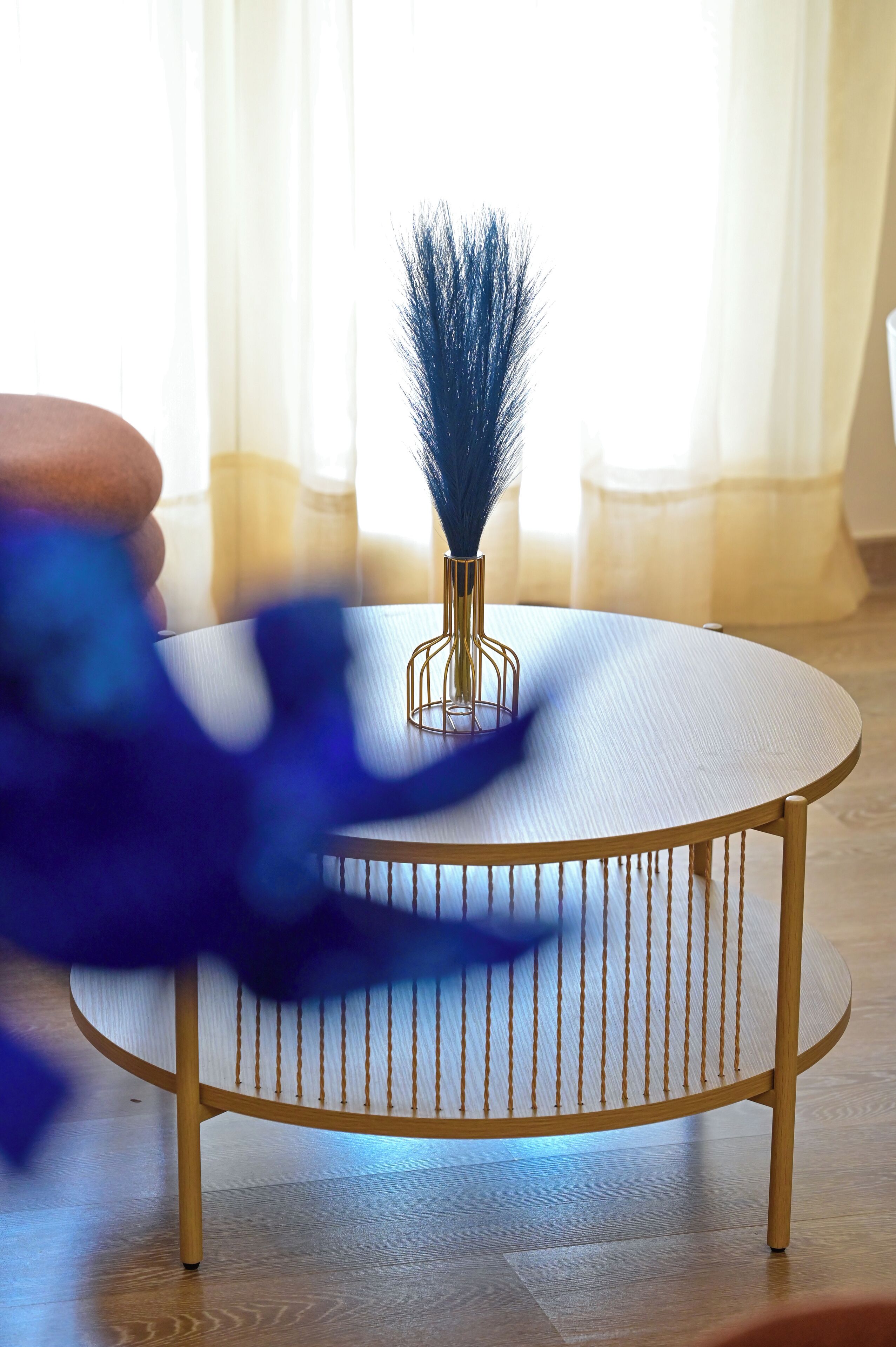 A round wooden table with a metal wireframe vase containing blue feathers, set on a wooden floor with a white tablecloth.