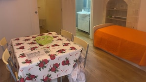 Een kamer met een tafel bedekt met een tafelkleed met kersenprint, een bed met een oranje deken, een stenen openhaard en een badkamer zichtbaar door een geopende deur.