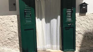 Een groene deur met een witte gordijn, een kleine brievenbus en een technische kast op een witte muur.