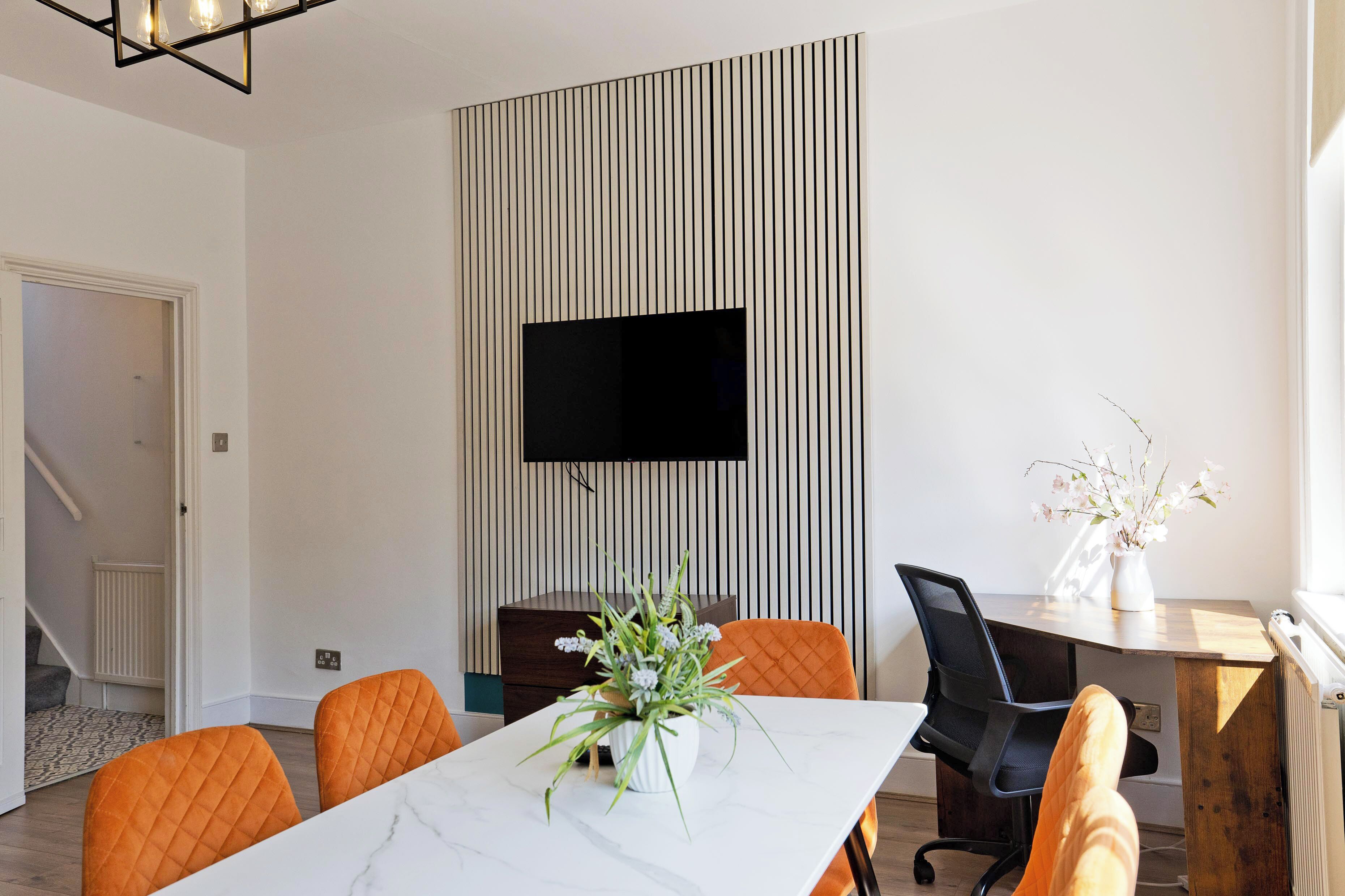 Een moderne eethoek met een witte marmeren tafel, oranje stoelen, een potplant, een zwarte bureaustoel, een houten bureau en een hangende televisie.