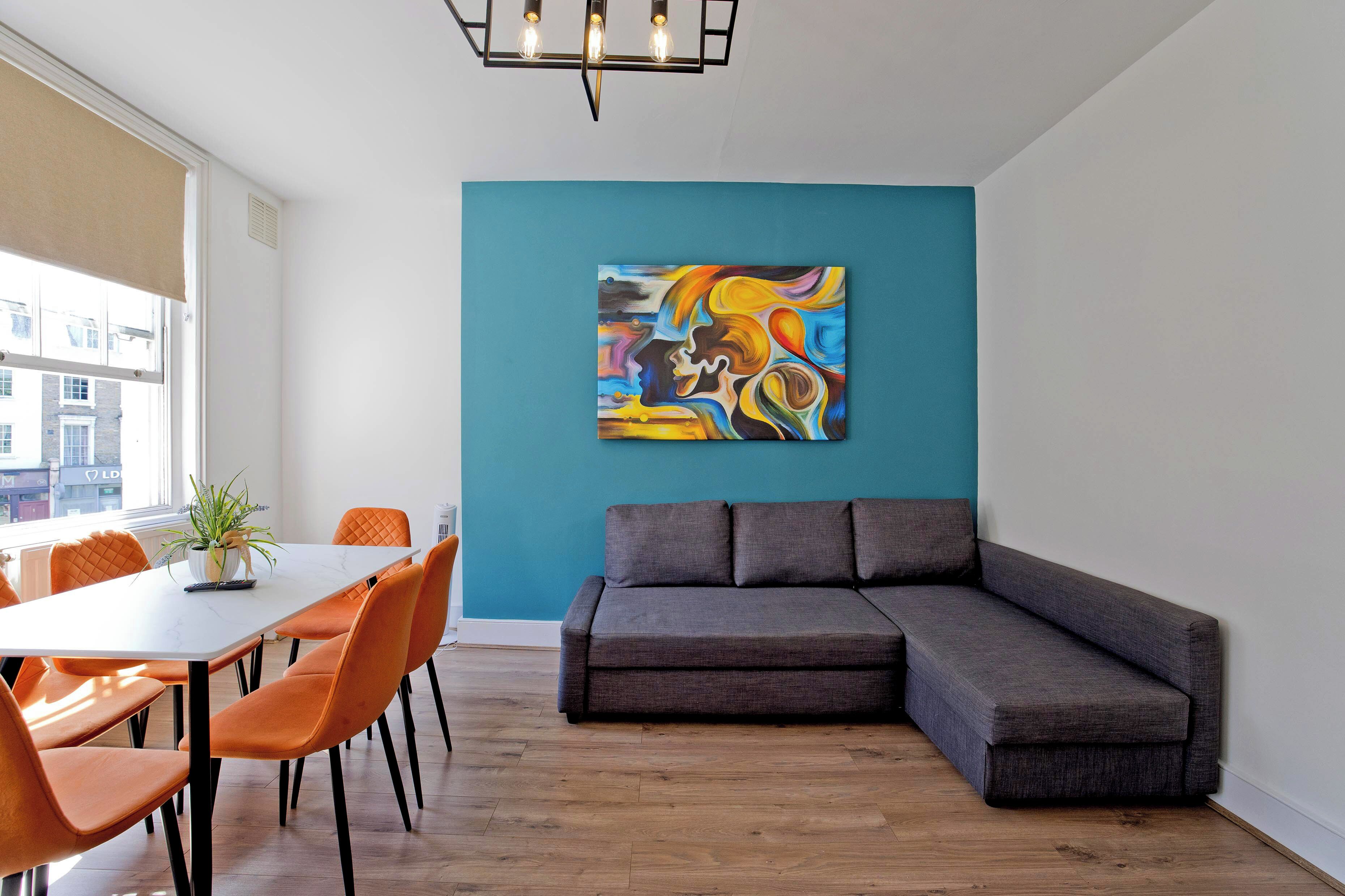 A modern living room with a gray sectional sofa, a white dining table with orange chairs, and a colorful abstract painting on the teal wall.