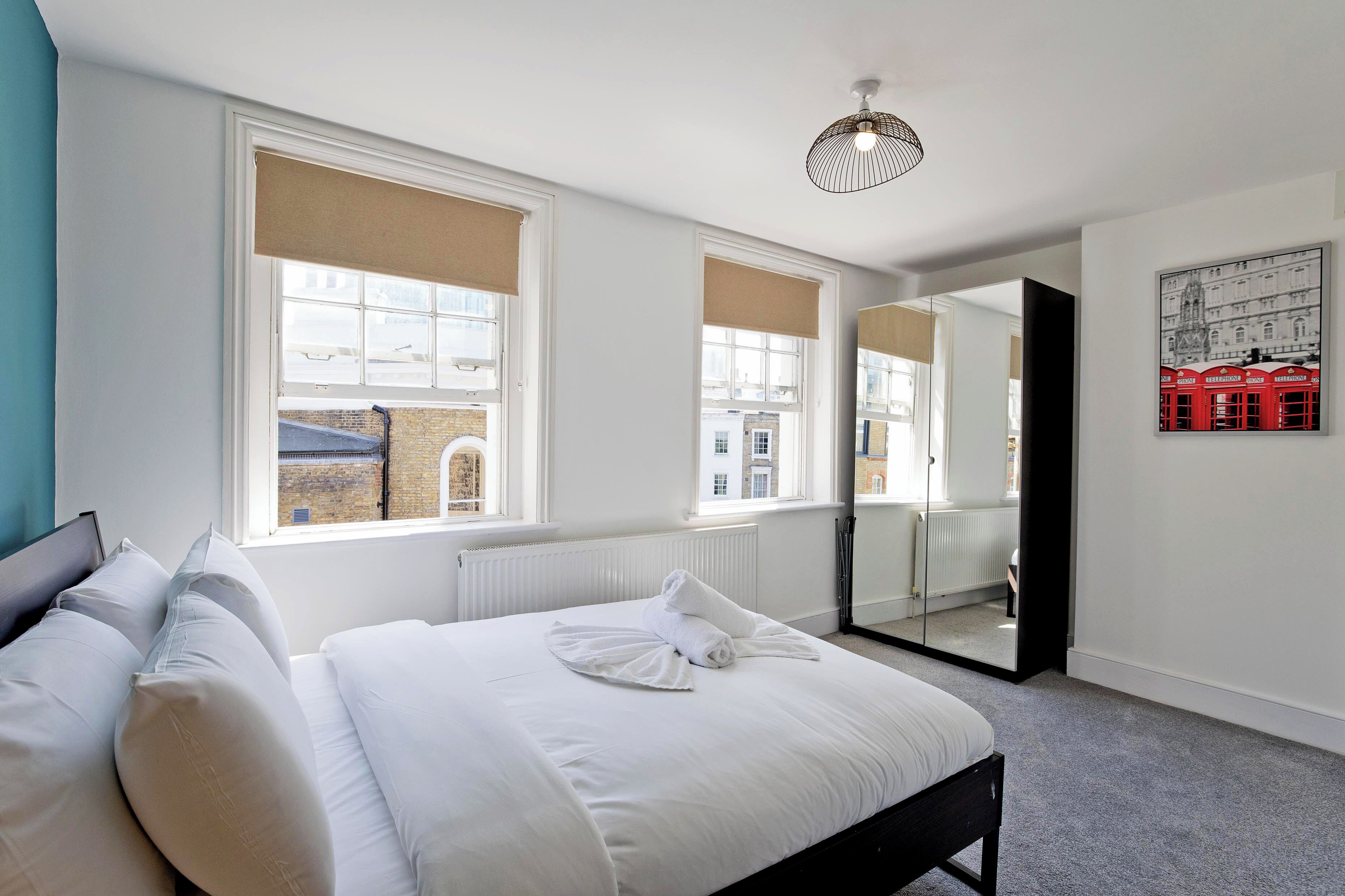 A bedroom with a large bed, two windows with blinds, a radiator, a mirror, and a wall-mounted light fixture.