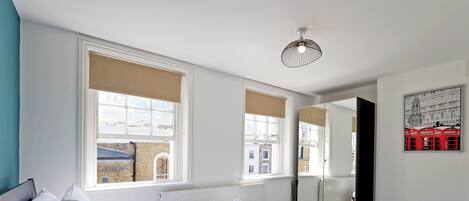 A bedroom with a large bed, two windows with blinds, a radiator, a mirror, and a wall-mounted light fixture.