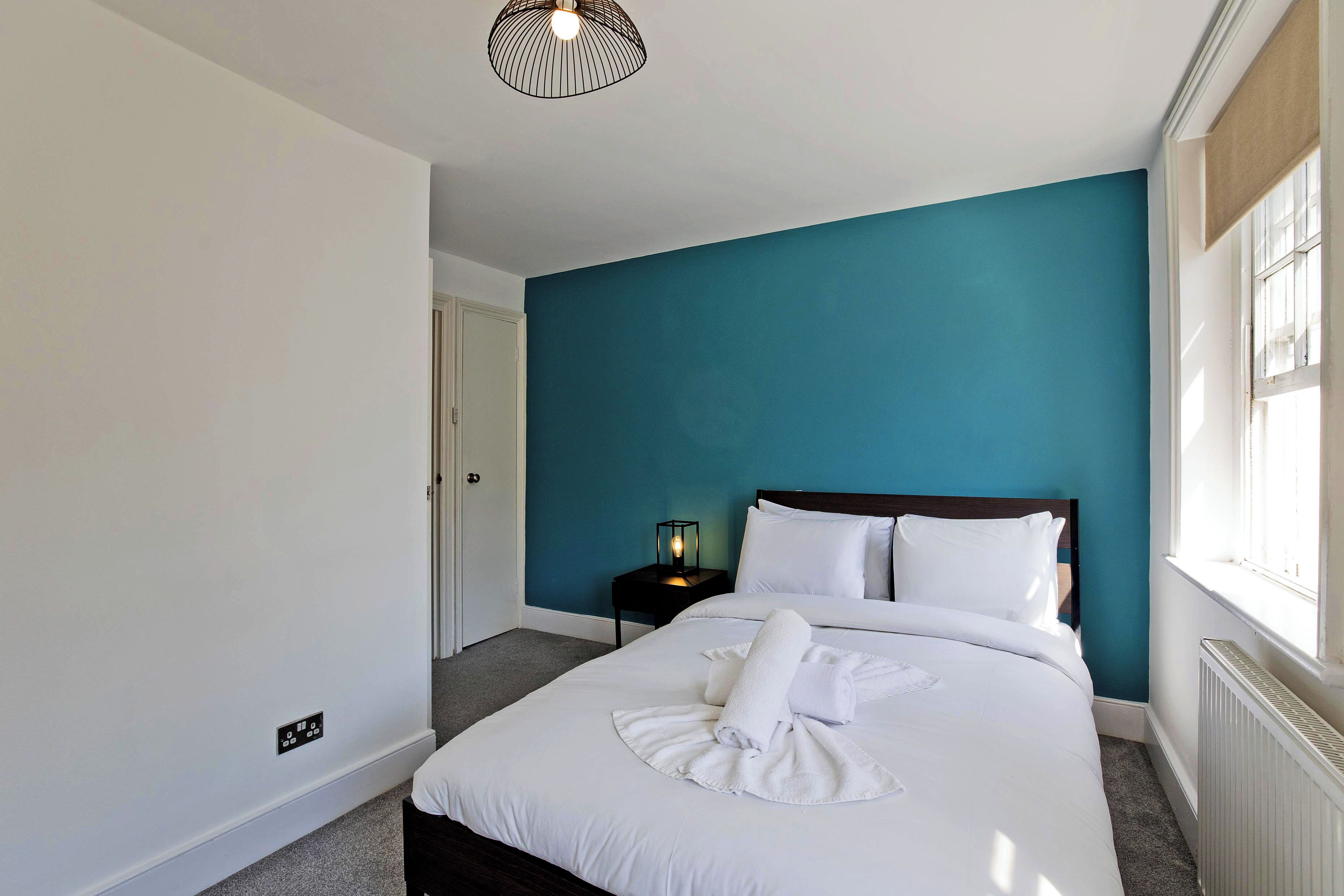 A neatly made bed with white linens and a towel, a nightstand with a lamp, and a window with blinds.
