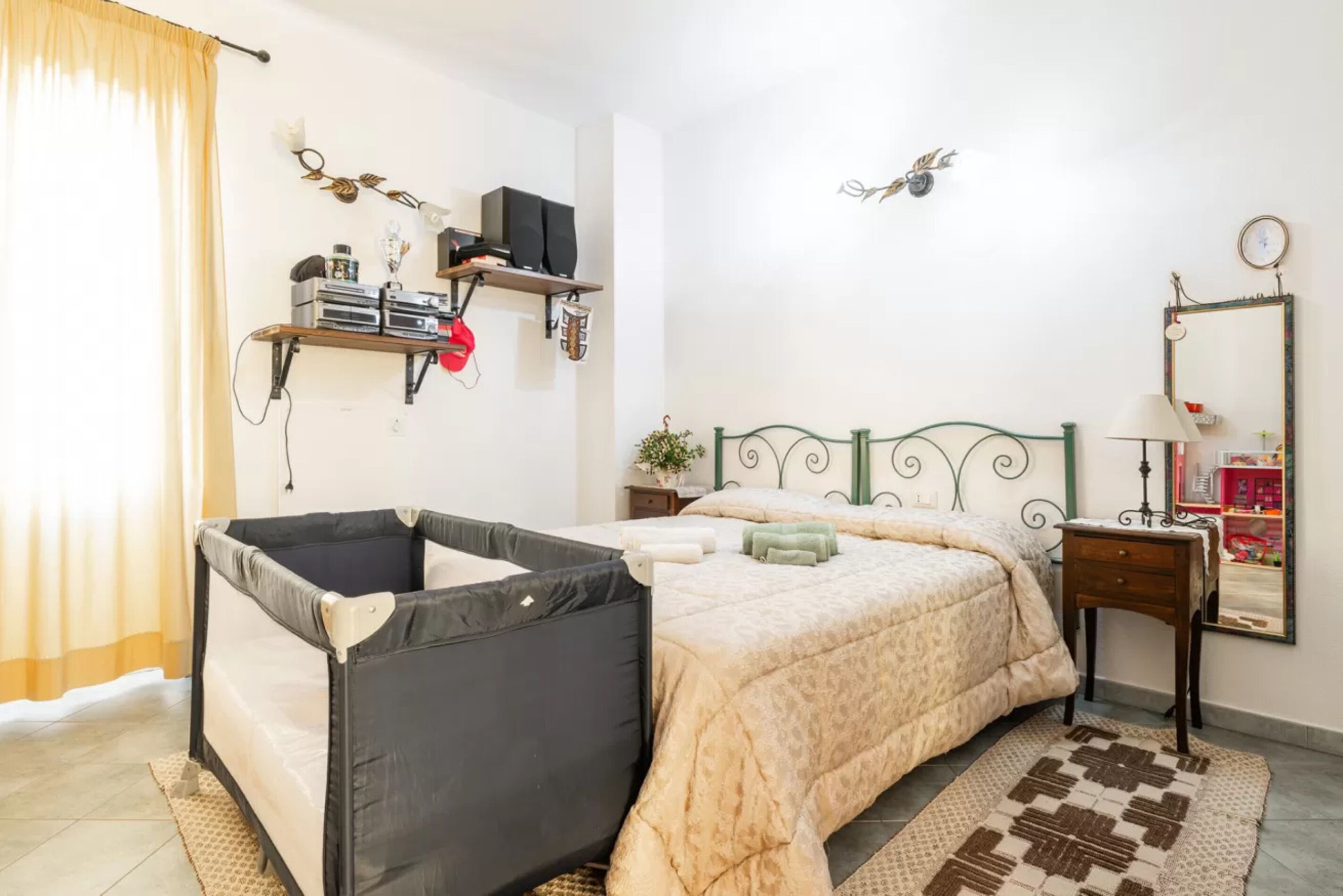 A bedroom with a bed, a small bedside table, a mirror, and a shelf with electronic devices.