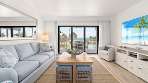 A modern living room with a sofa, coffee table, and TV, overlooking a balcony with a pool and palm trees. - Direct Beachfront and View | Walk-Out to Sand | Pool + Pickleball + Tennis | (Captiva)