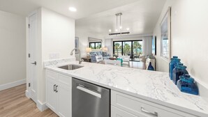 A modern kitchen with a marble countertop, stainless steel sink, and dishwasher. There is a dining area with a table and chairs, and a view of the outdoors through large windows. - Direct Beachfront and View | Walk-Out to Sand | Pool + Pickleball + Tennis | (Captiva)