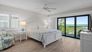 A bedroom with a bed, a chair, a nightstand, a ceiling fan, and a balcony with a view of the ocean.