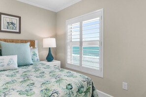 Un dormitorio con cama, mesita de noche, lámpara, ventana con vista al océano y un cuadro en la pared.