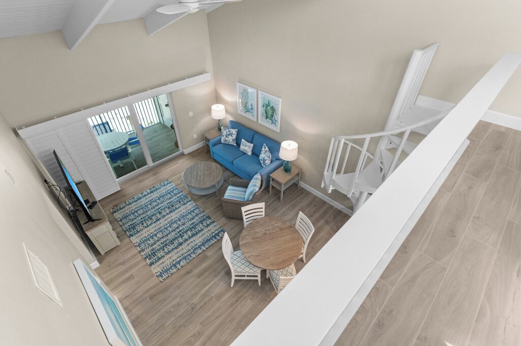 A spacious living area with a blue sofa, round coffee table, and white chairs. There is a large window with blinds, a ceiling fan, and a staircase leading to another level.