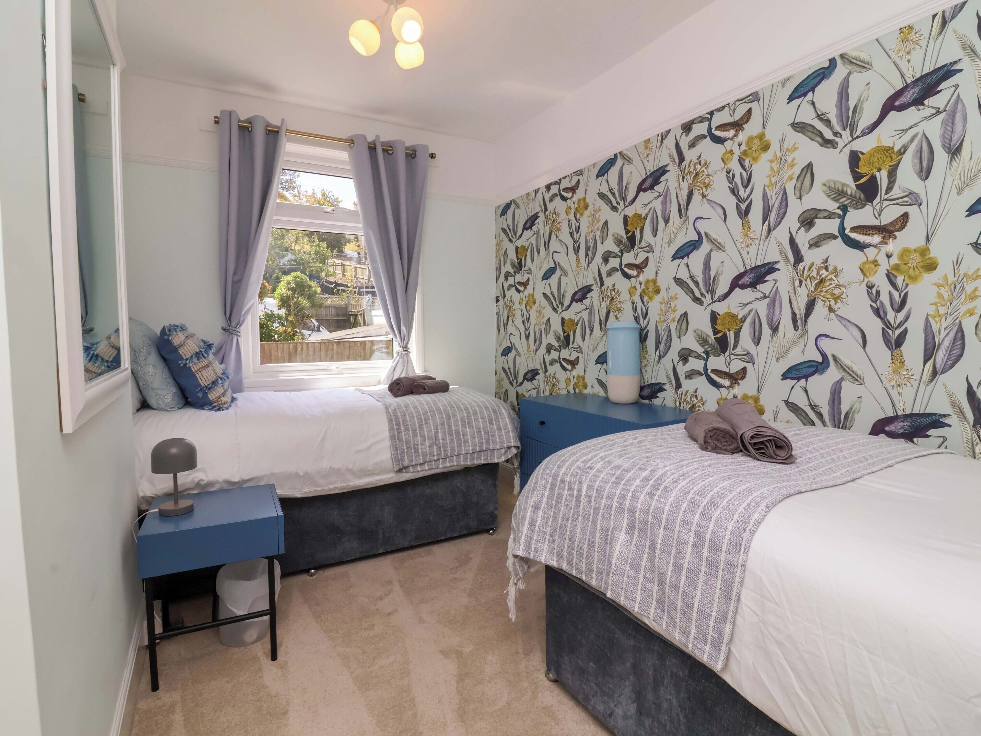 A bedroom with two beds, a window with curtains, a desk with a lamp, and a wall with a floral pattern.