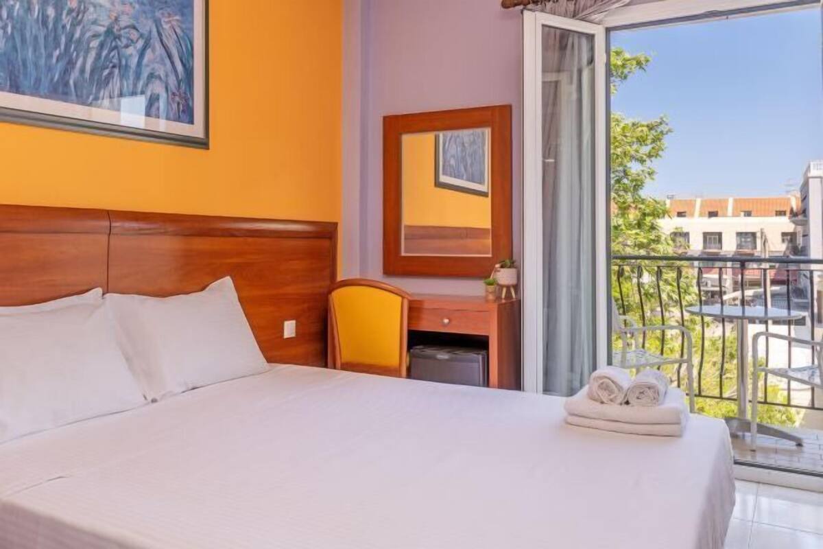 Ein Hotelzimmer mit einem Bett, einem Schreibtisch, einem Stuhl, einem kleinen Tisch mit Handtüchern, einem Spiegel und Blick ins Freie.