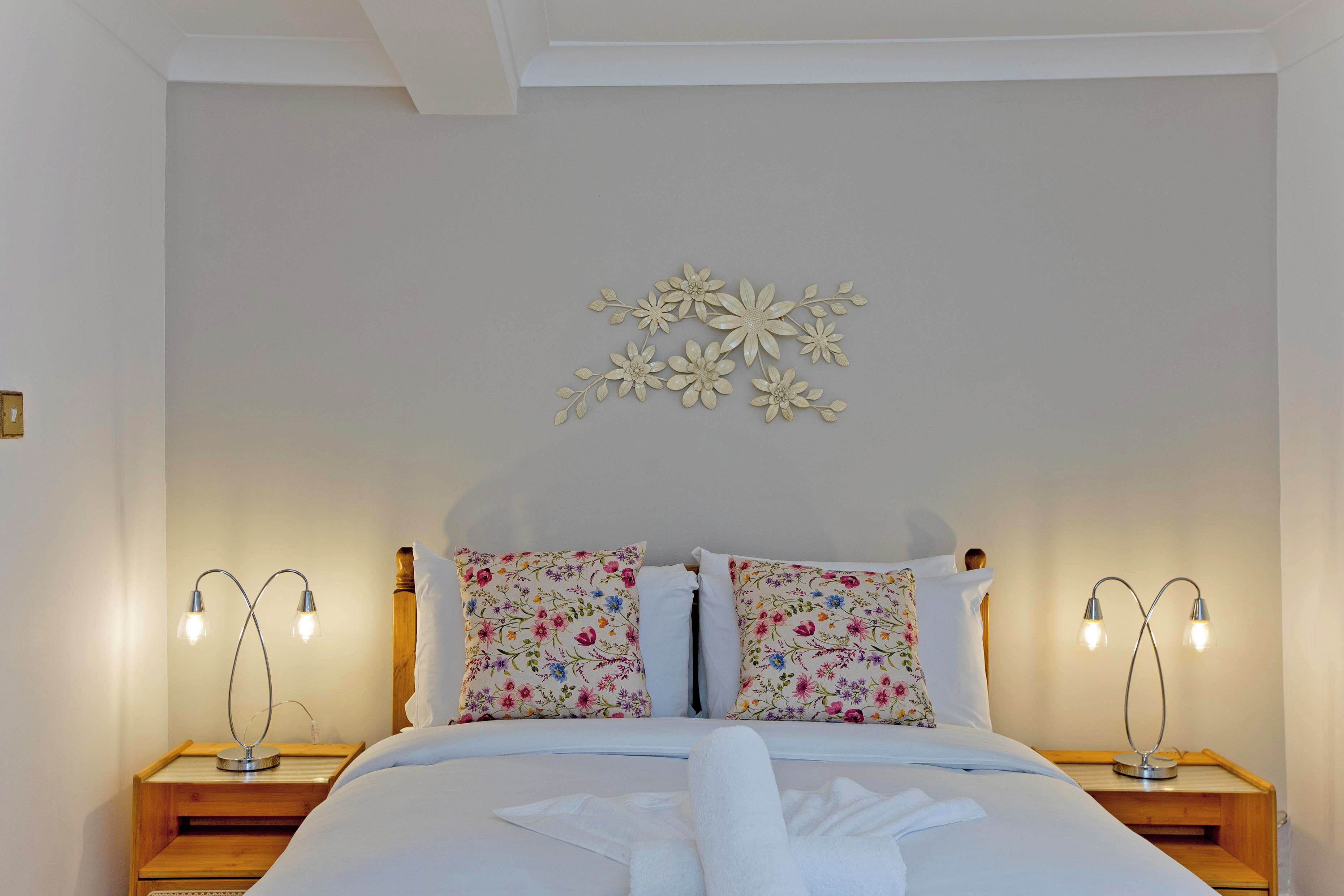 Una cama bien tendida con almohadas estampadas, dos lámparas de noche y una decoración floral en la pared.