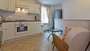 A kitchen with white cabinets, a toaster, an oven, and a refrigerator. There is a chair with a floral cushion and a table with a glass top.