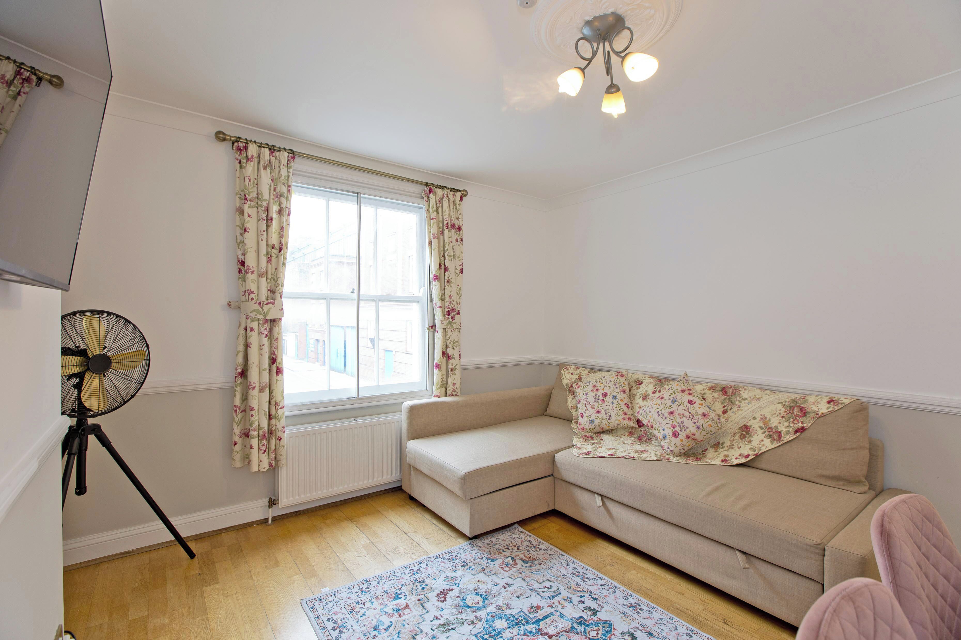 Un salon chaleureux avec un canapé beige, un plaid à motifs floraux, un ventilateur sur pied et une fenêtre ornée de rideaux à fleurs.