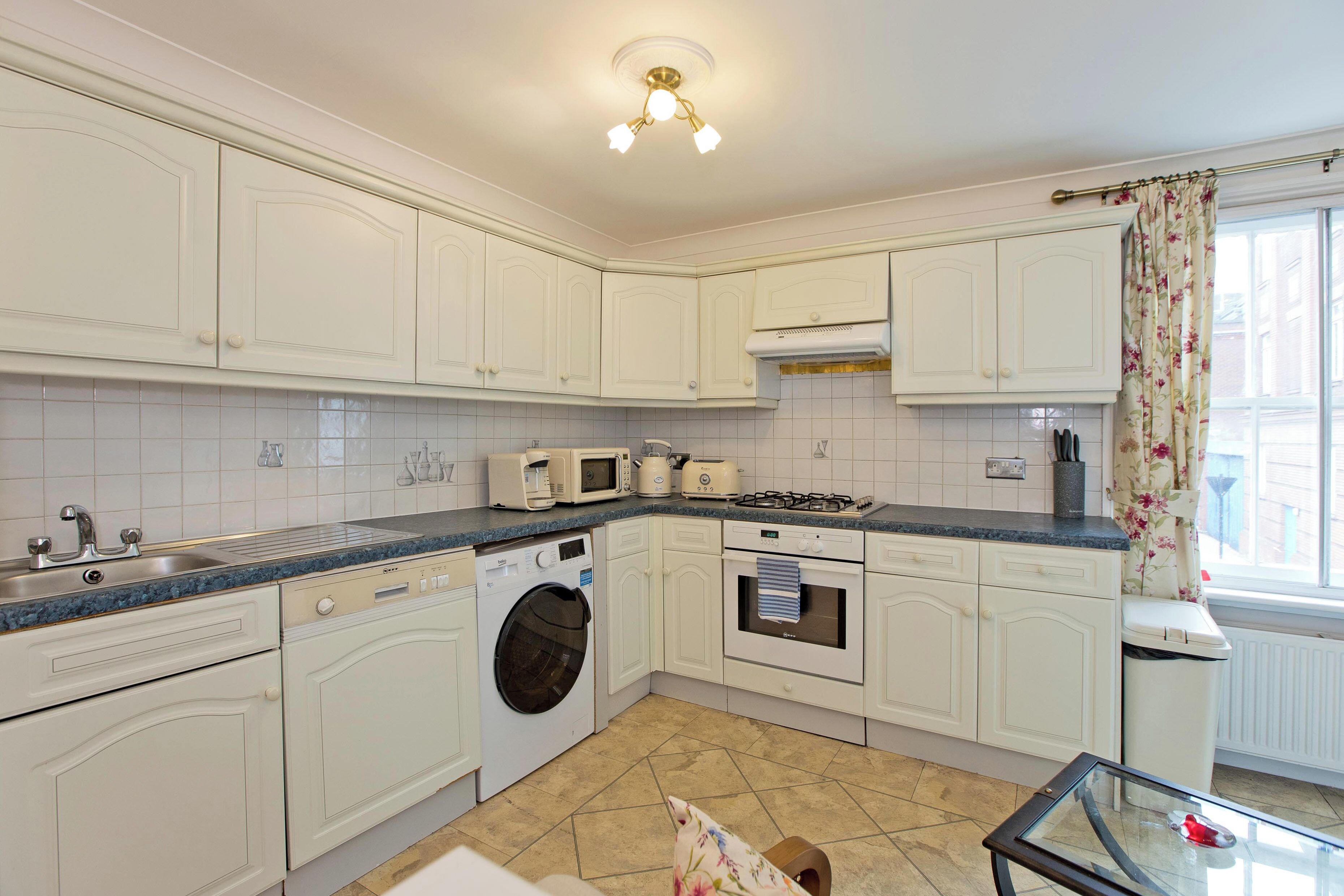 A kitchen with white cabinets, a washing machine, a microwave, a toaster, and a glass table.
