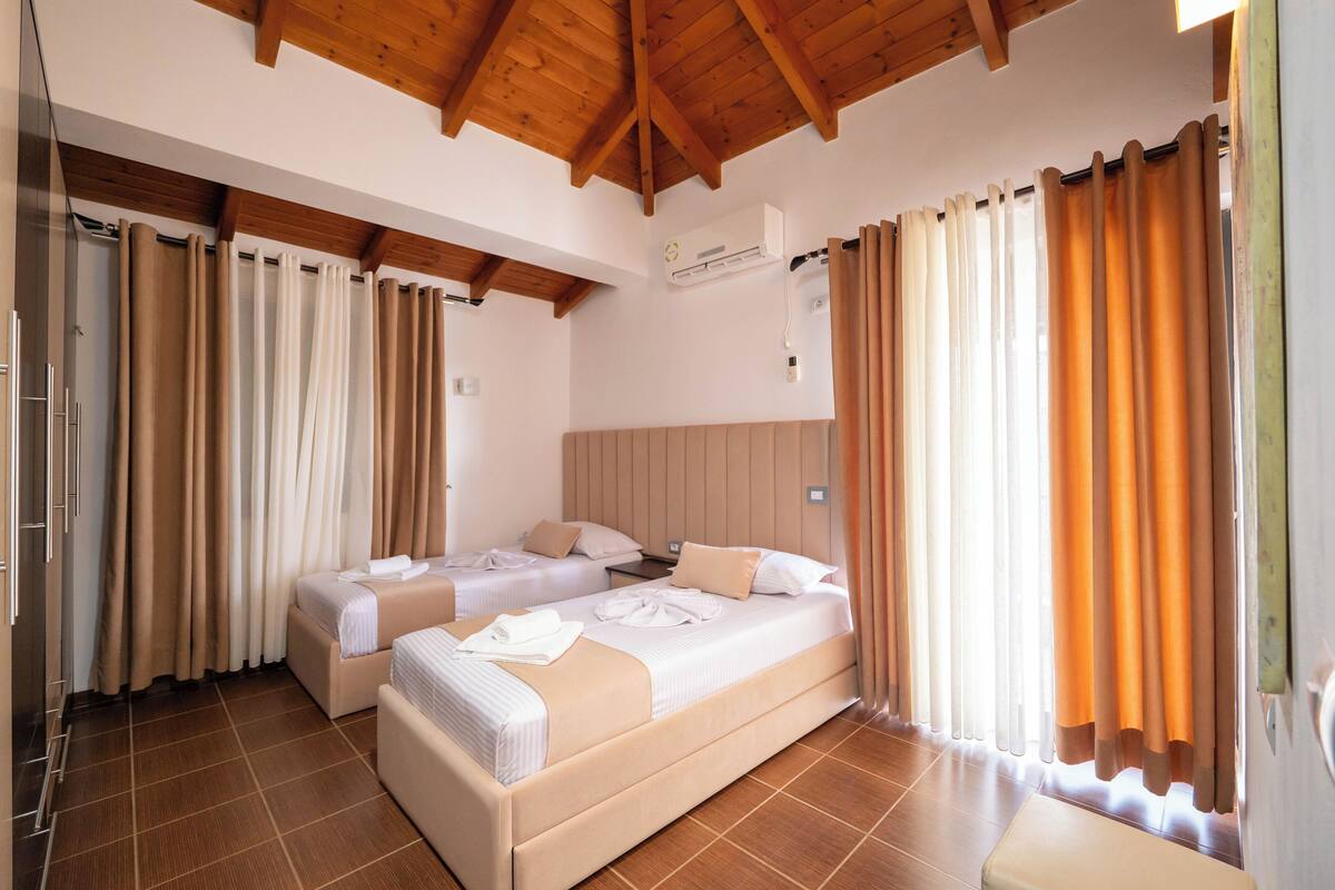 Habitación de hotel con dos camas, techo de madera y ventana con cortinas naranjas.