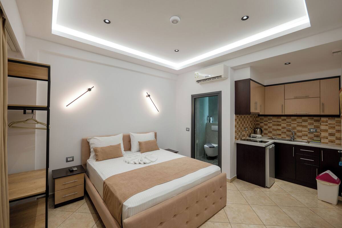Una habitación de hotel moderna con cama, mesitas de noche, armario, baño y kitchenette.