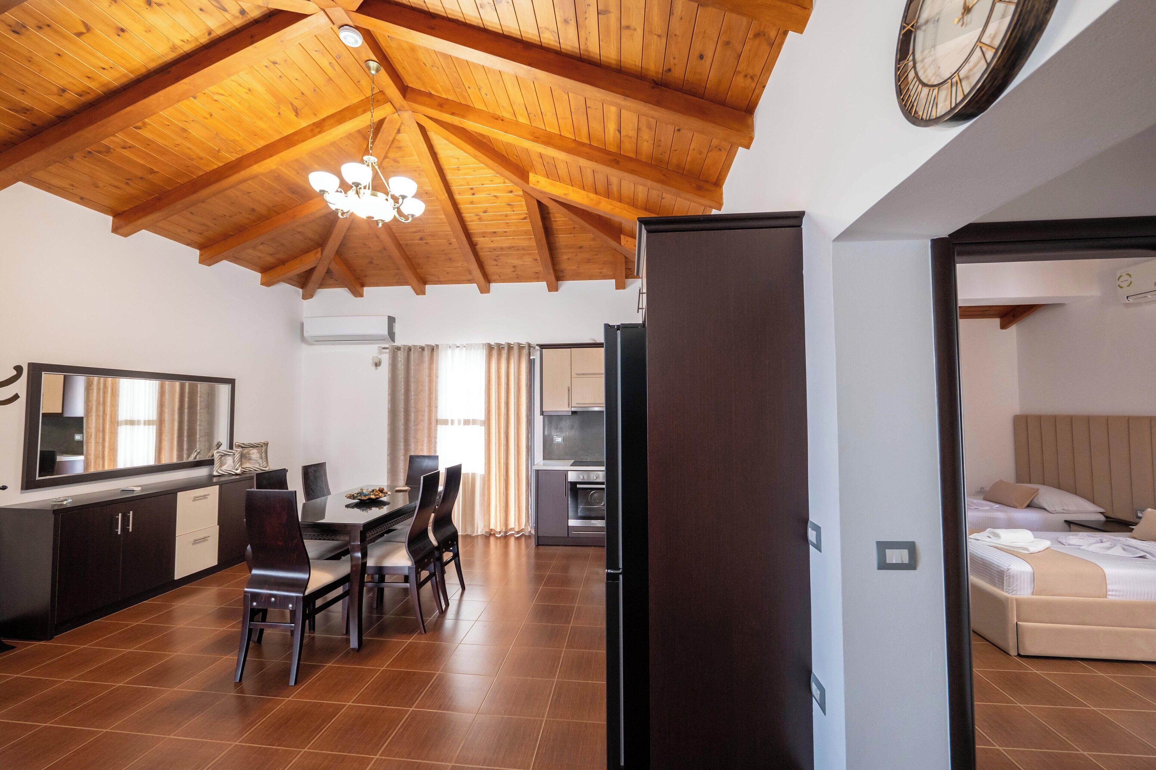 A modern kitchen with wooden beams, a dining area with chairs and a table, and a bedroom with a bed and headboard.