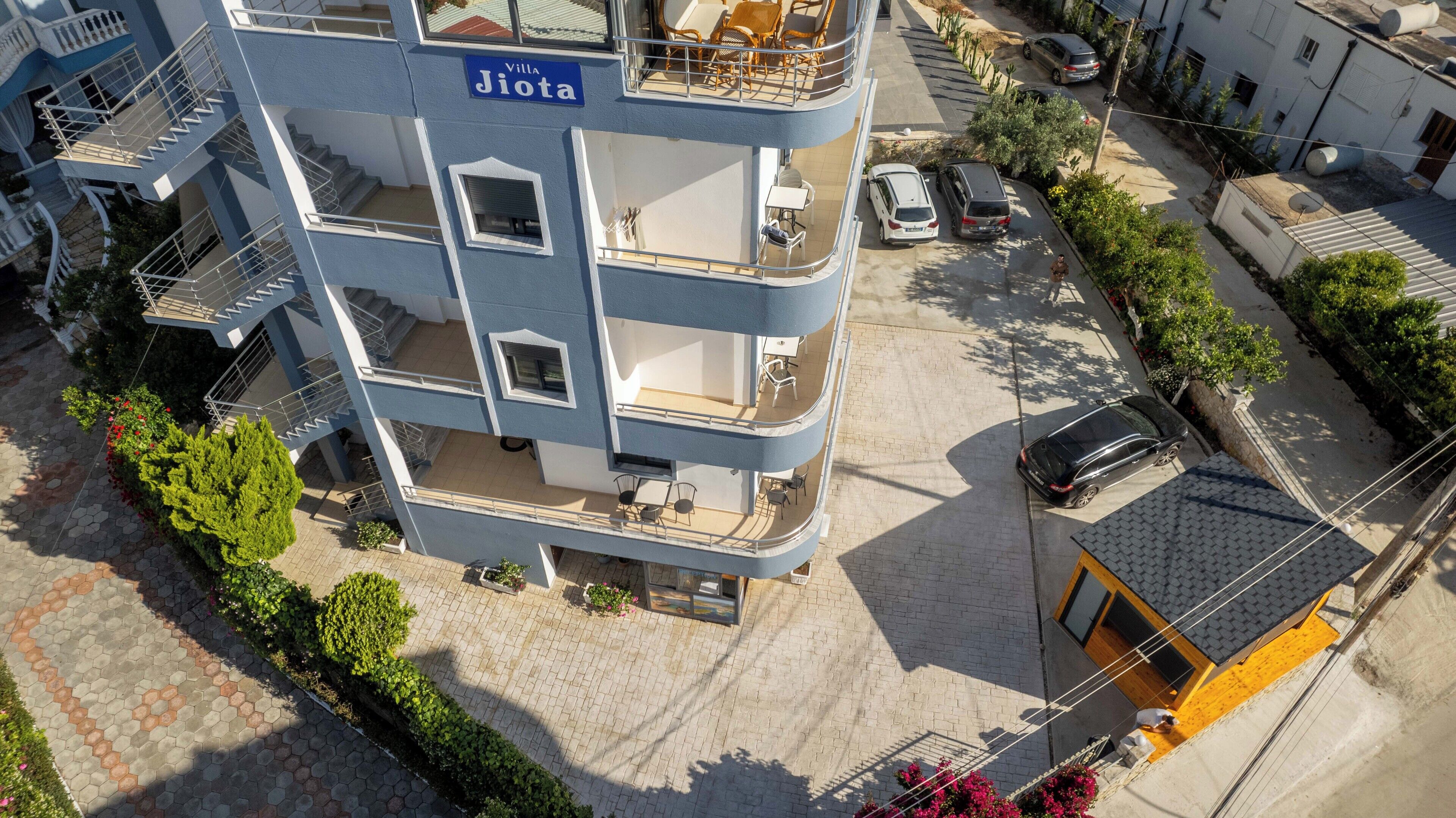 A multi-story building with balconies, a sign reading "Villa Jiota," and a parking area with cars.