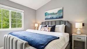 A bedroom with a large bed, a nightstand, a window with a view of trees, and a painting on the wall.