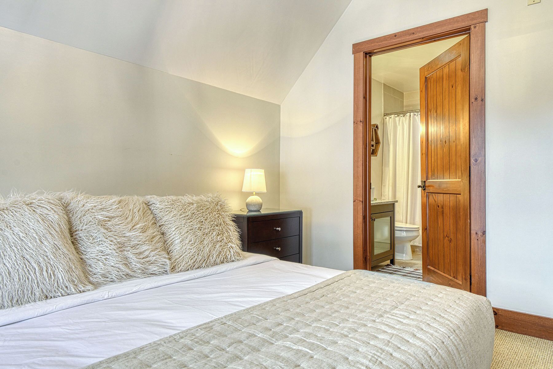 A bedroom with a bed, a nightstand, a lamp, and a wooden door leading to a bathroom.