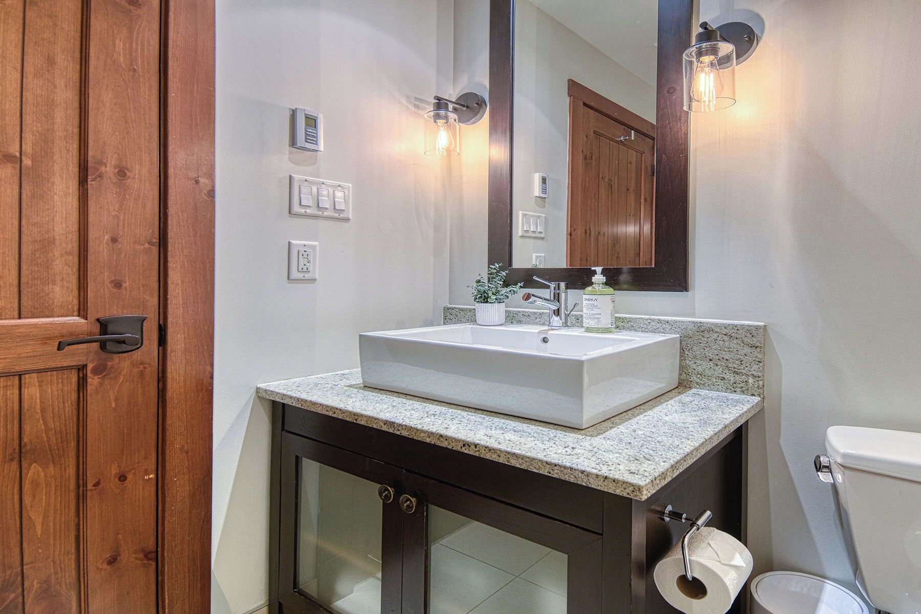 A bathroom with a wooden door, a white sink with a granite countertop, a mirror above the sink, and a toilet with a paper towel holder.