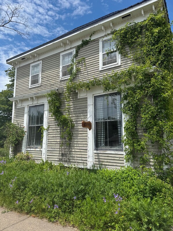 Une maison à deux étages, ornée d’une enseigne de style victorien, entourée d’une végétation luxuriante et de plantes en fleurs.