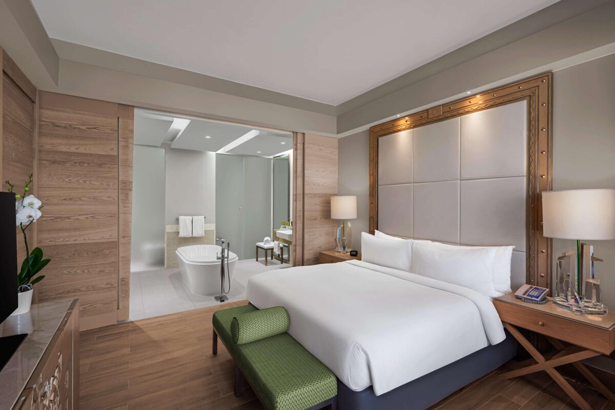 A hotel room with a large bed, a bedside table, a green bench, a bathroom with a bathtub, and a TV.