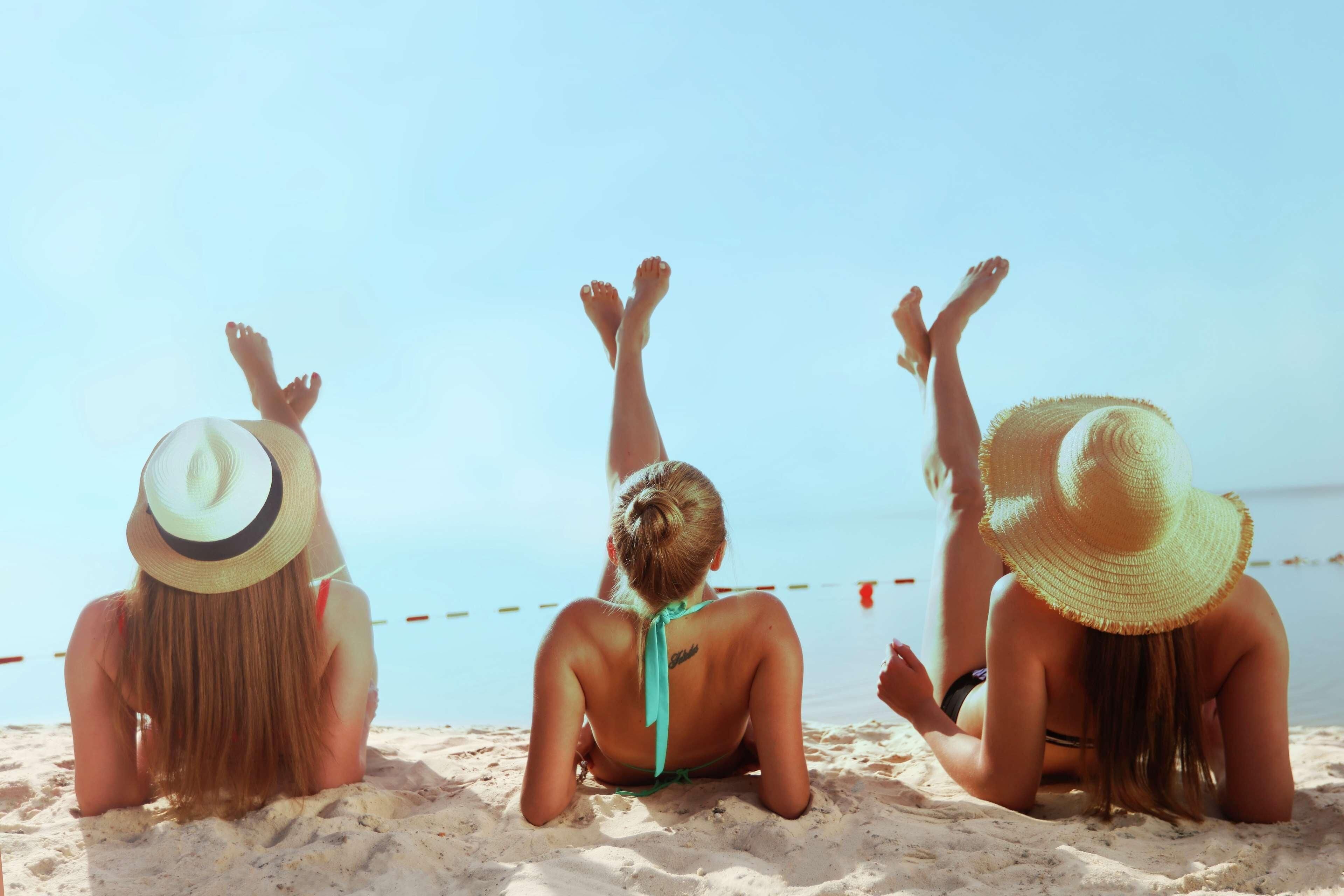 Three individuals sitting on a sandy beach with their backs to the camera, wearing hats and swimwear.