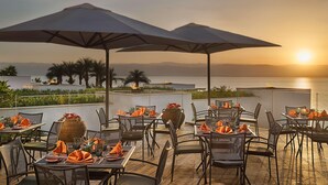 An outdoor dining area with tables, chairs, umbrellas, and a view of the sea at sunset. - Hilton Dead Sea Resort & Spa (Sweimeh)