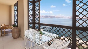 A balcony with a glass table and white wicker chairs, overlooking a body of water and distant land. - Hilton Dead Sea Resort & Spa (Sweimeh)