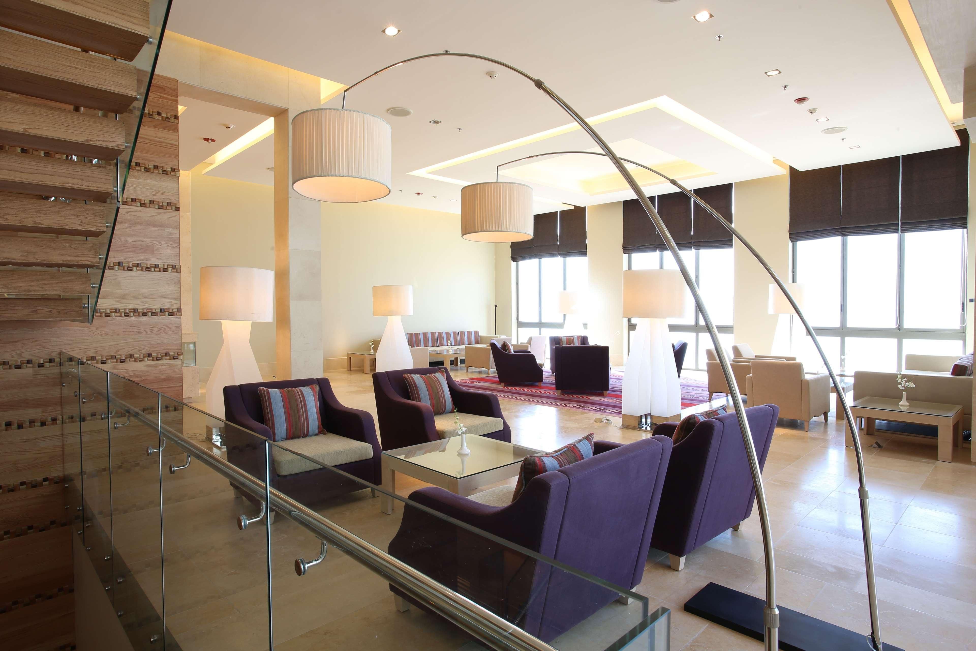 Een moderne hotel lobby met een glazen trap, een zithoek met banken en stoelen, en grote ramen voor natuurlijk lichtinval.