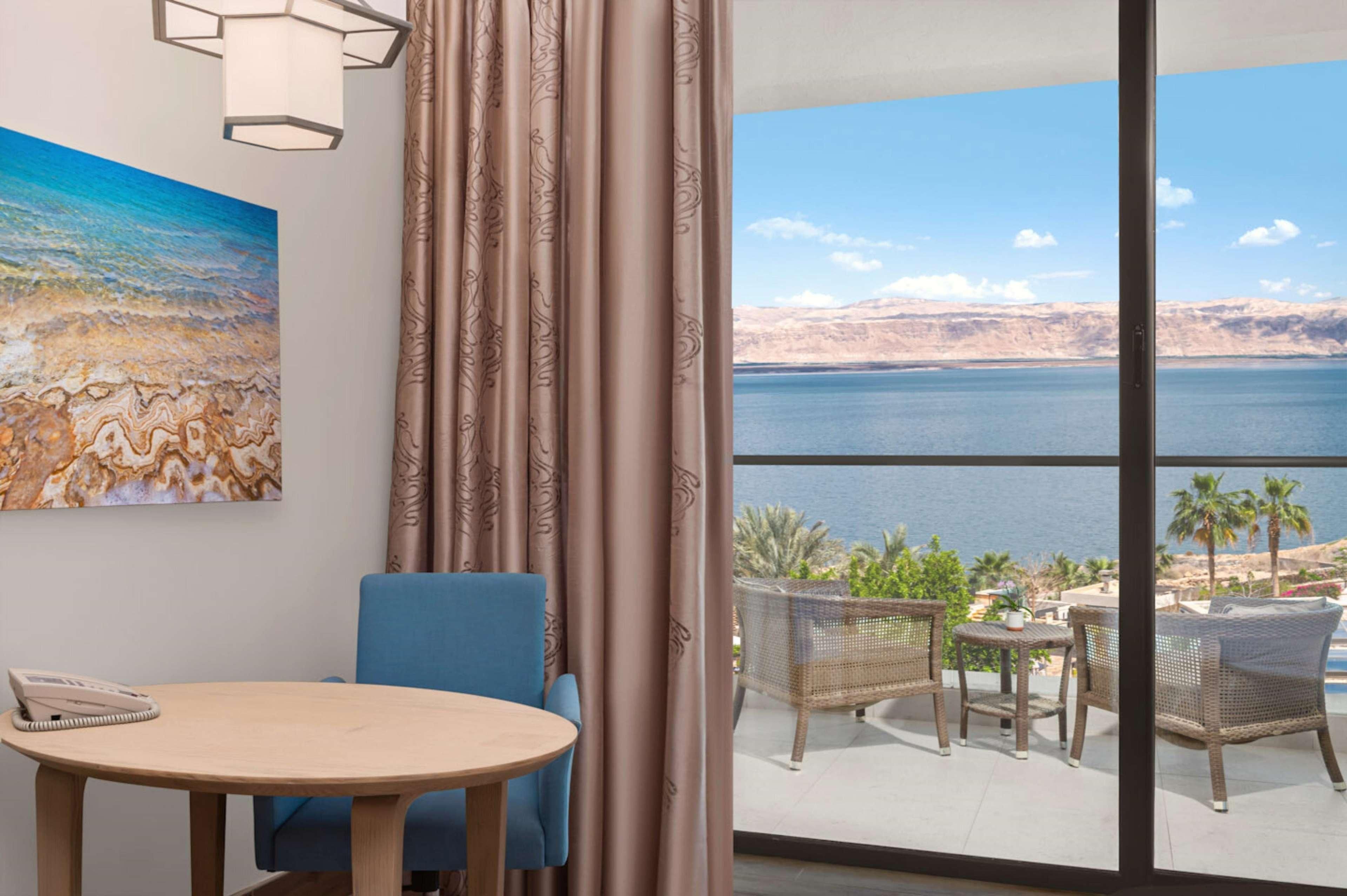 A room with a view of a large body of water, a painting of a beach scene, a round table with a chair, and a balcony with wicker furniture.