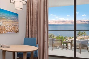 A room with a view of a large body of water, a painting of a beach scene, a round table with a chair, and a balcony with wicker furniture. - Hilton Dead Sea Resort & Spa (Sweimeh)