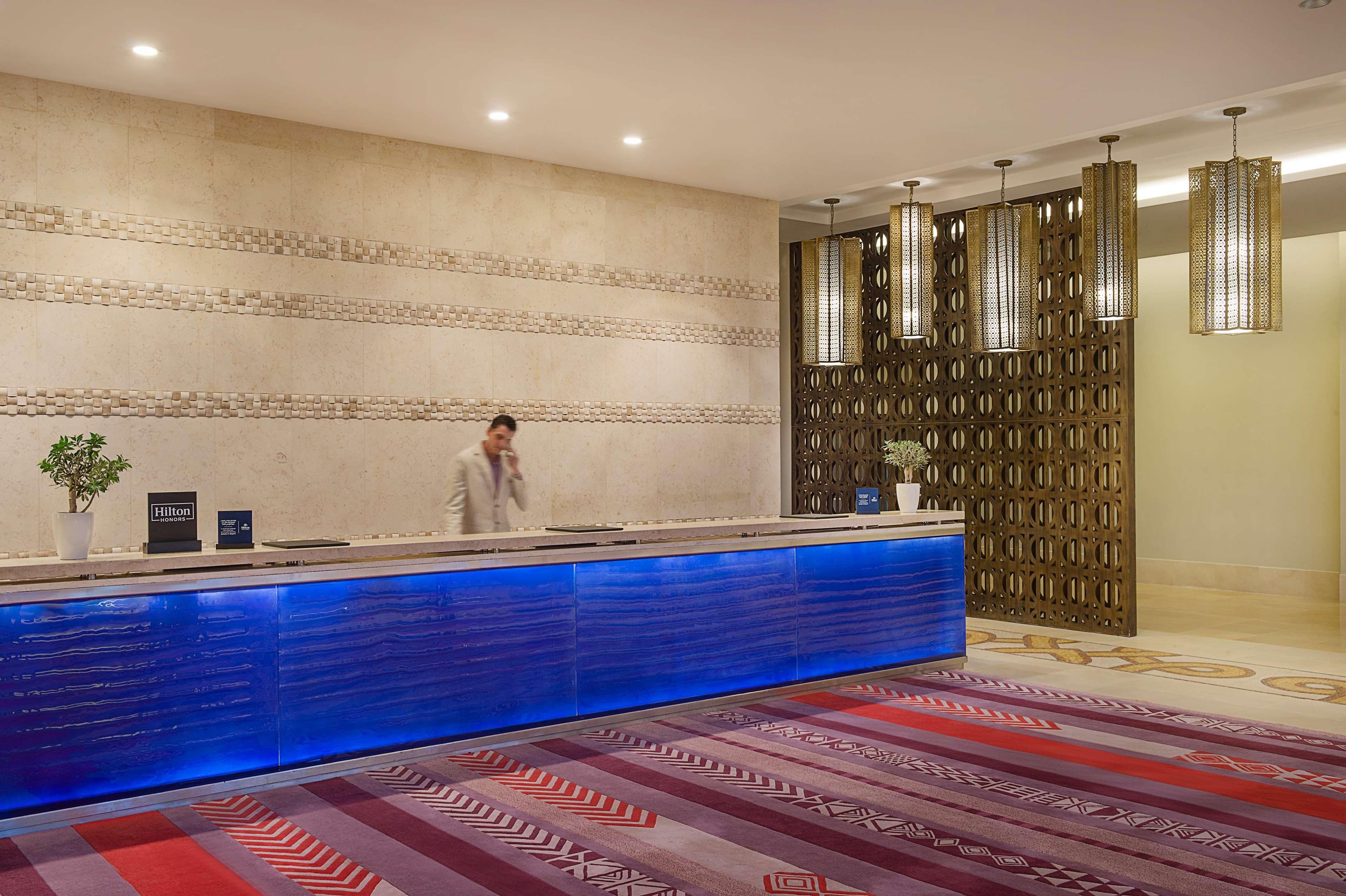A hotel reception desk with a blue illuminated counter, a potted plant, and a decorative wall with a geometric pattern.
