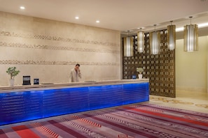 A hotel reception desk with a blue illuminated counter, a potted plant, and a decorative wall with a geometric pattern. - Hilton Dead Sea Resort & Spa (Sweimeh)