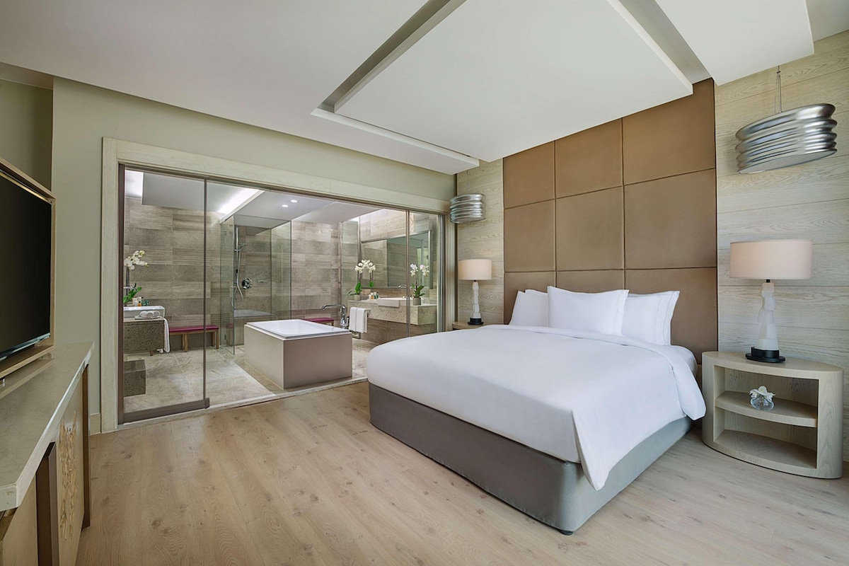 A modern hotel room with a large bed, a glass-enclosed shower area, a flat-screen TV, and a wooden floor.