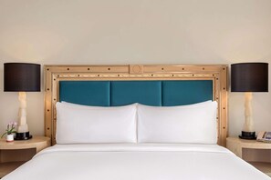 Un lit bien fait, avec du linge de lit blanc, une tête de lit recouverte d’un tissu turquoise et deux lampes de chevet posées sur des tables de chevet assorties.
