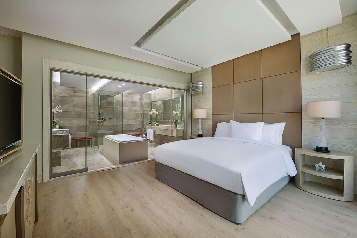 A modern hotel room with a large bed, a glass-enclosed shower area, a flat-screen TV, and a wooden floor.