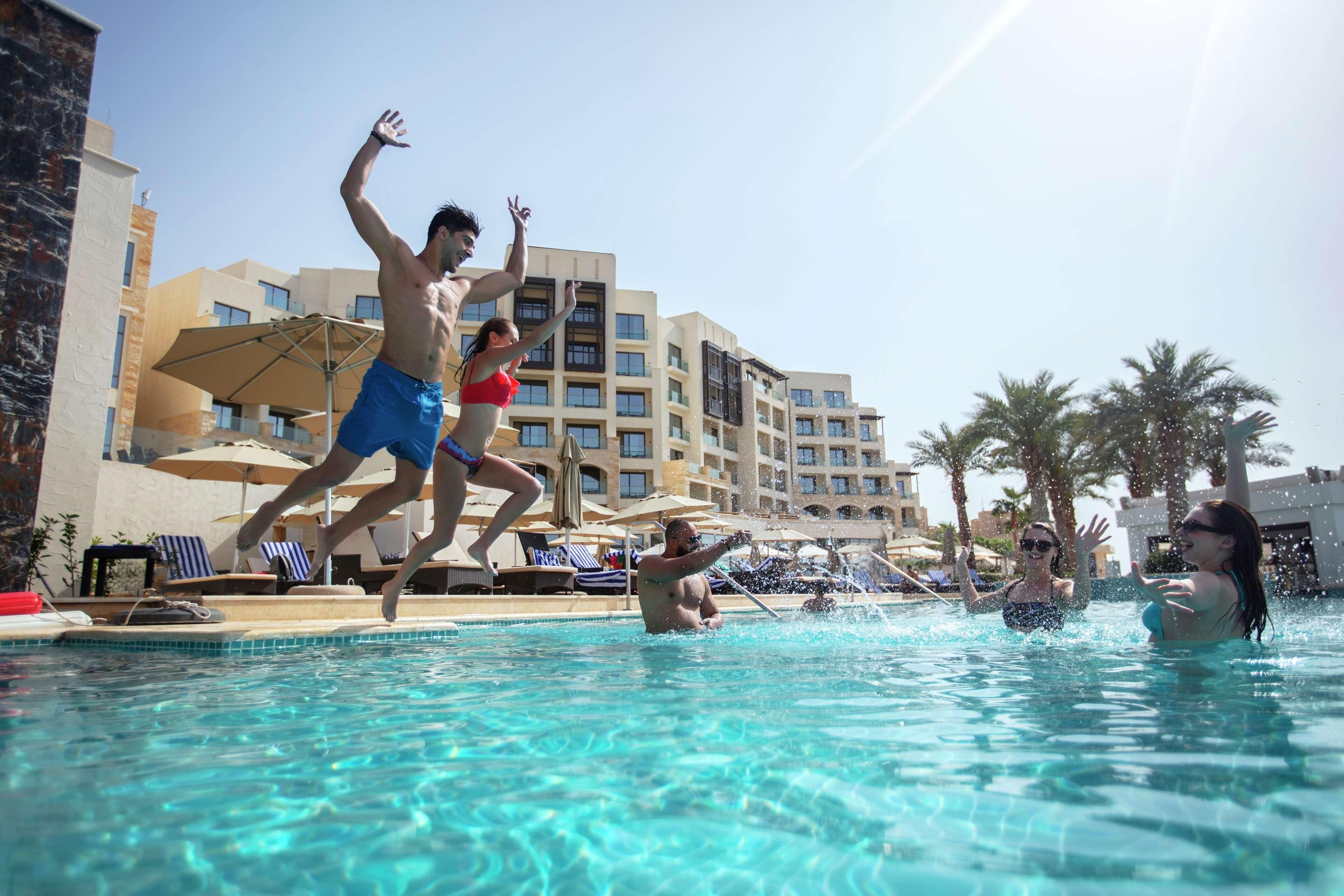 A swimming pool with people jumping, lounge chairs, umbrellas, and a hotel building in the background.