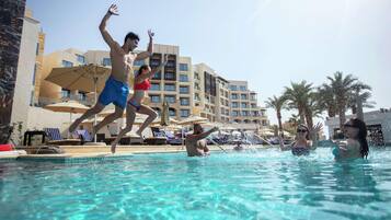 A swimming pool with people jumping, lounge chairs, umbrellas, and a hotel building in the background.